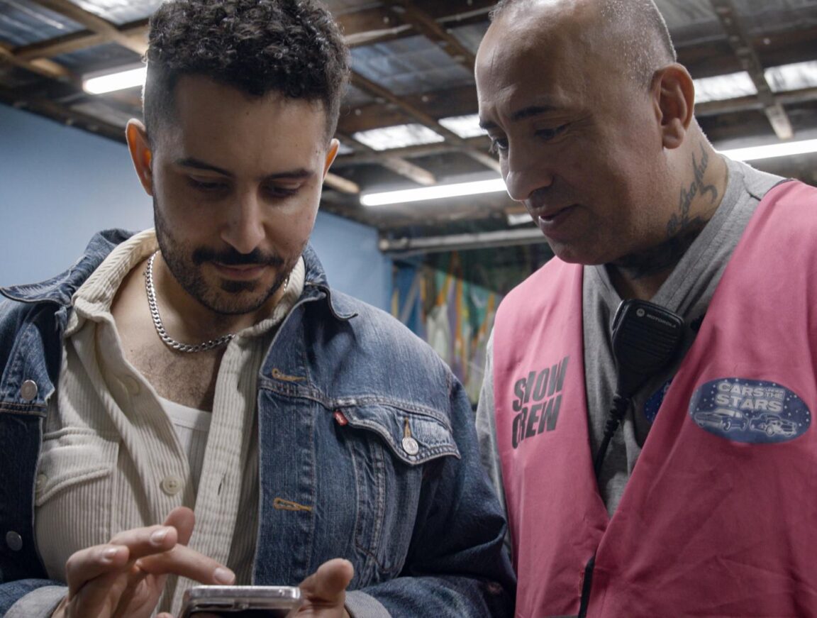 Two men, one in a pink high-vis vest and the other in a denim jacket, looking intently over a smart phone.
