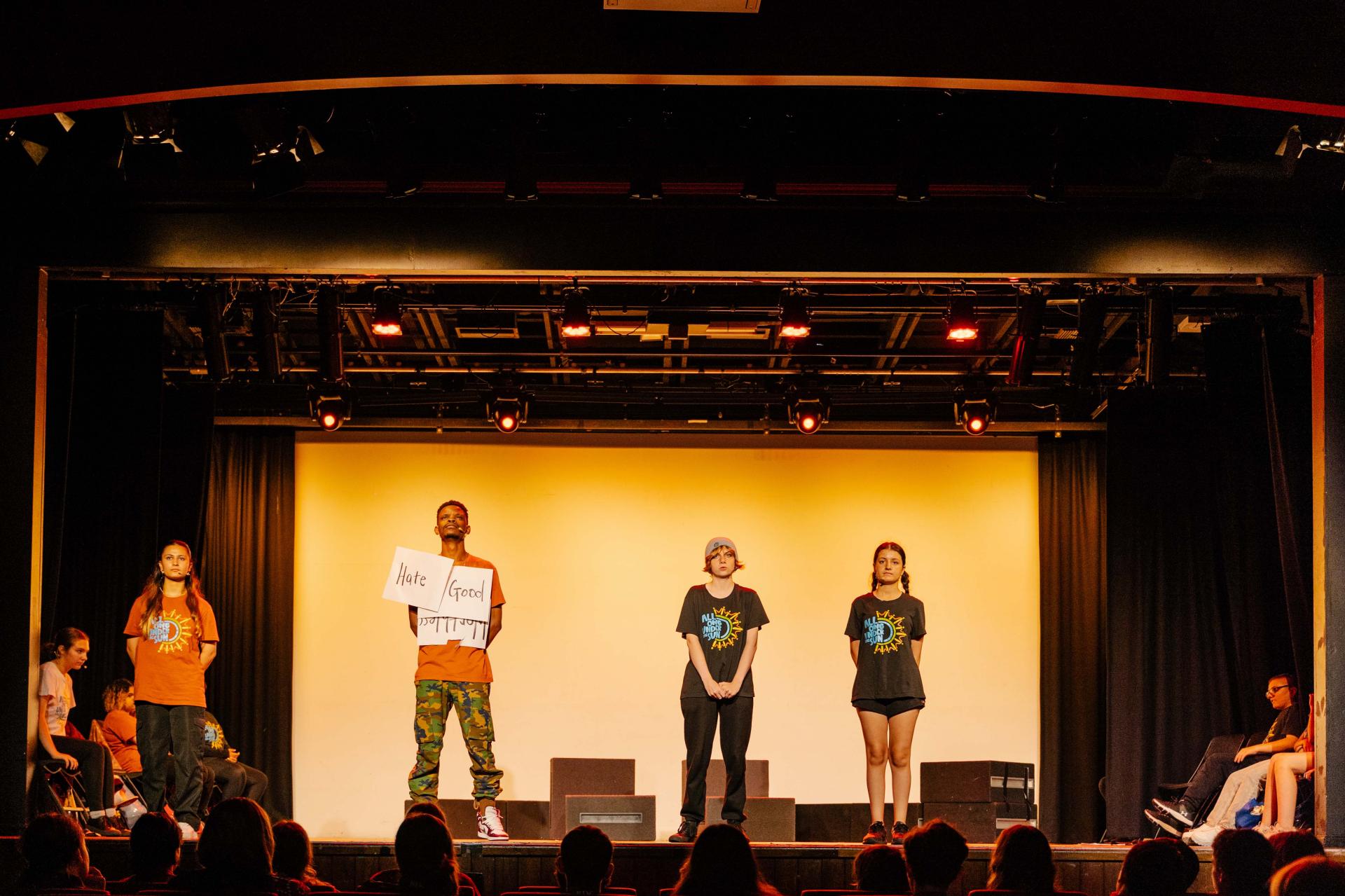Four people stand on a stage as part of a performance, one has posters on his shirt saying the words 'good' and 'hate'.