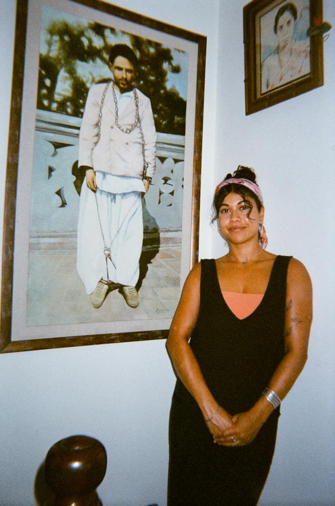 A woman in a black dress stands in front of a large, framed photograph of a man in white and chains
