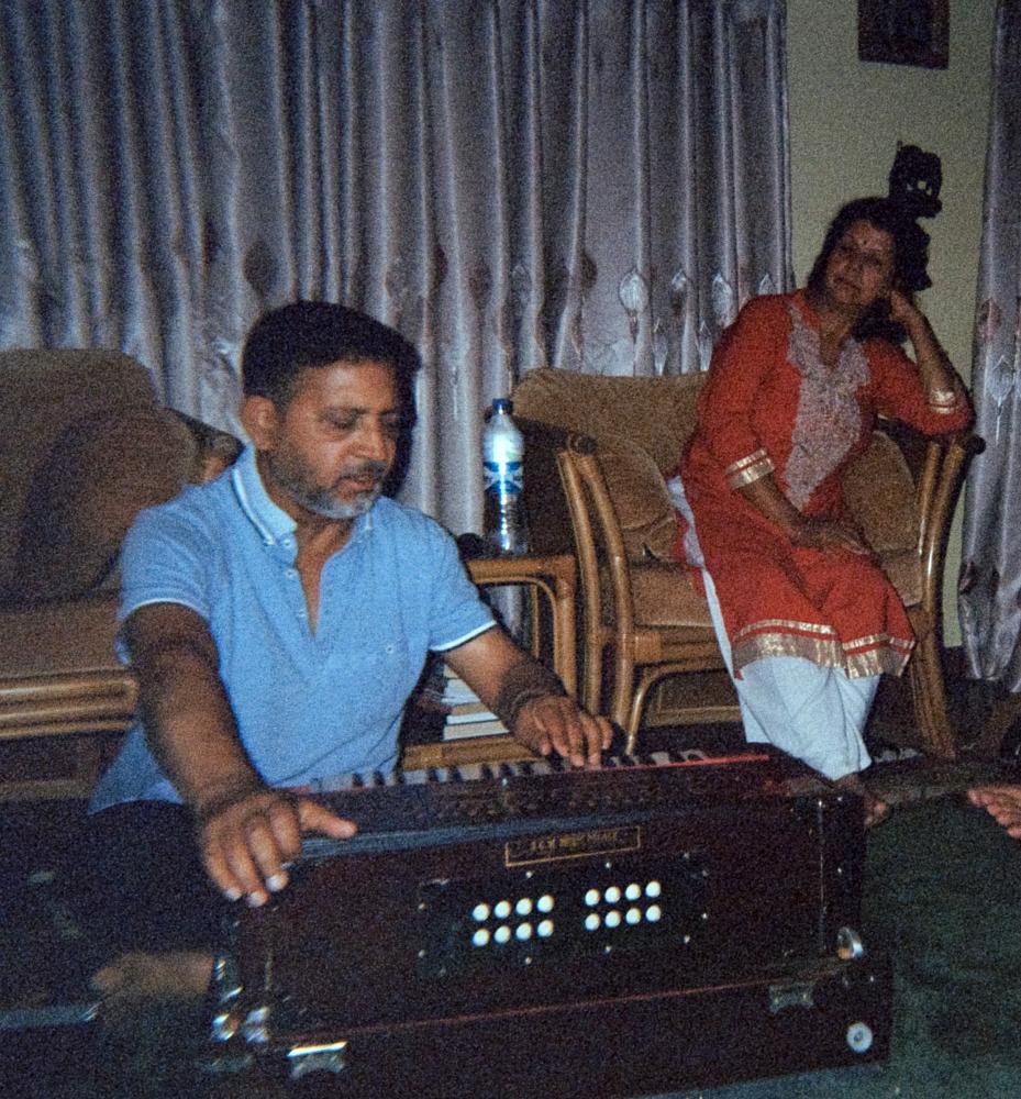 A man wearing a blue shirt sits on the floor playing with a music instrument, a harmonium, as a women in red traditional dress watches from a nearby chair