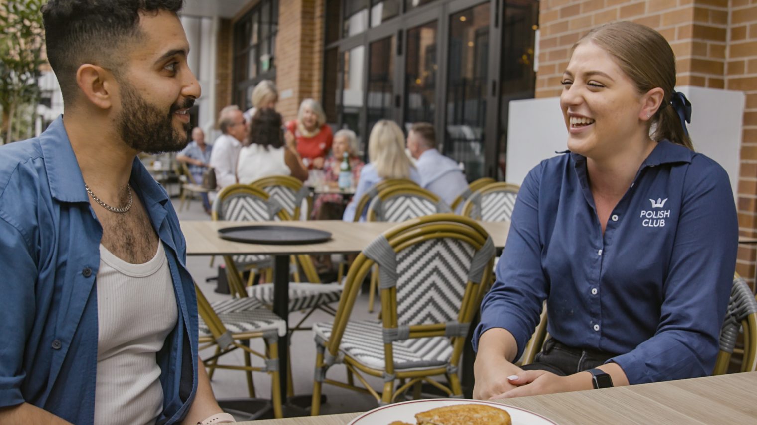 A man in an open blue shirt and white singlet and woman wearing a navy blue uniform with Polish Club embroidered, seated at a table in discussion