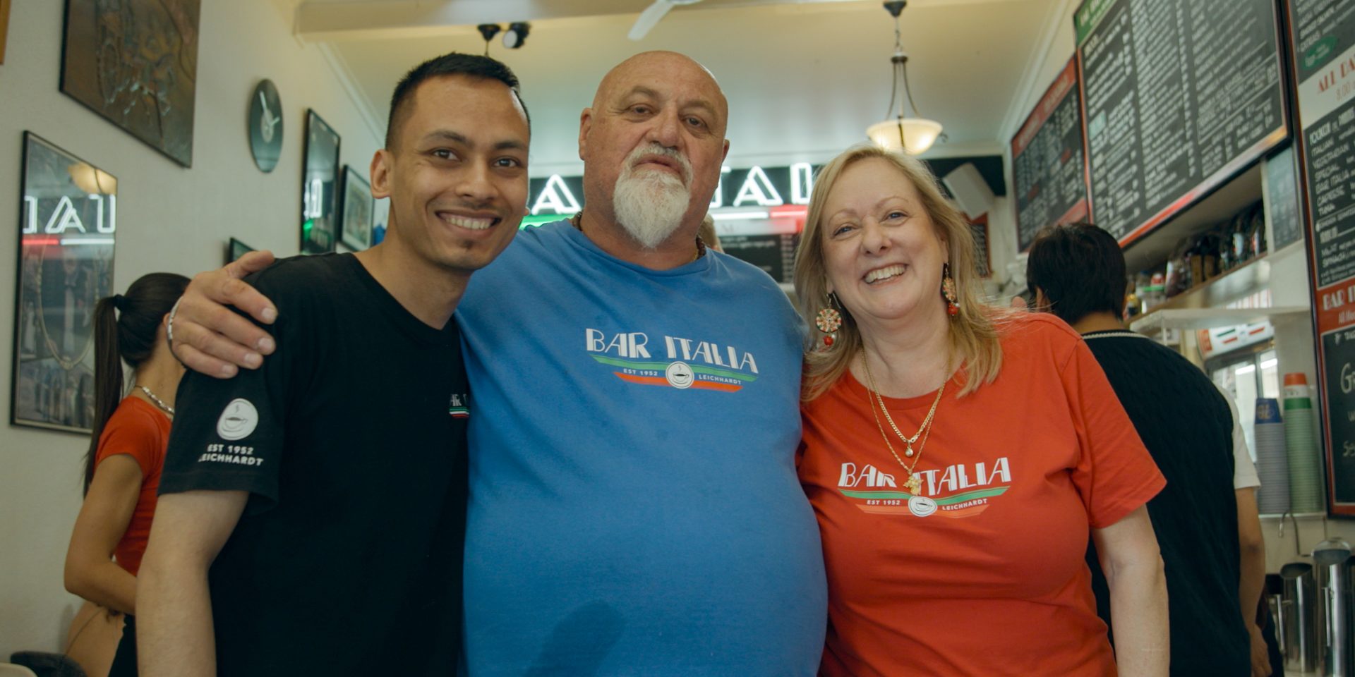 Two men and a woman in grouped together, wearing a black, a blue and a red shirt with the logo for Bar Italia restaurant, standing in said restaurant