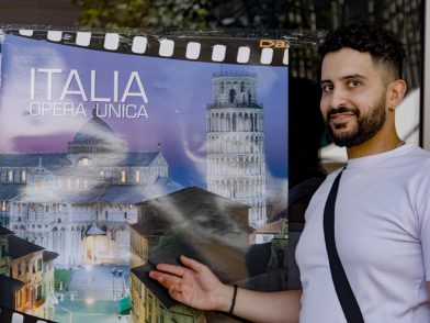 A man in a white t-shirt standing in front of a poster featuring the Pisa Cathedral and Tower of Pisa and the words "Italia Opera Unica"