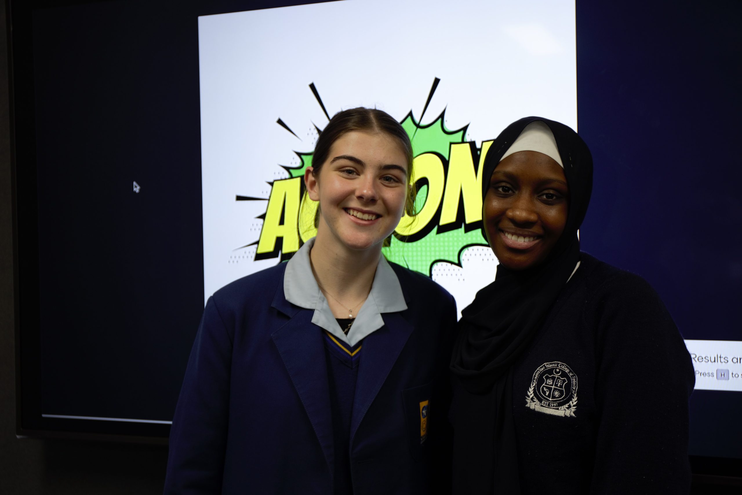 This image shows two smiling young women standing together in front of a screen that displays the word "ACTION!" in comic book-style text. The person on the left is wearing a school uniform with a light blue collar, and the person on the right is wearing a dark hijab and a matching school jumper with a logo on it. Both seem to be from different schools, given their distinct uniforms.